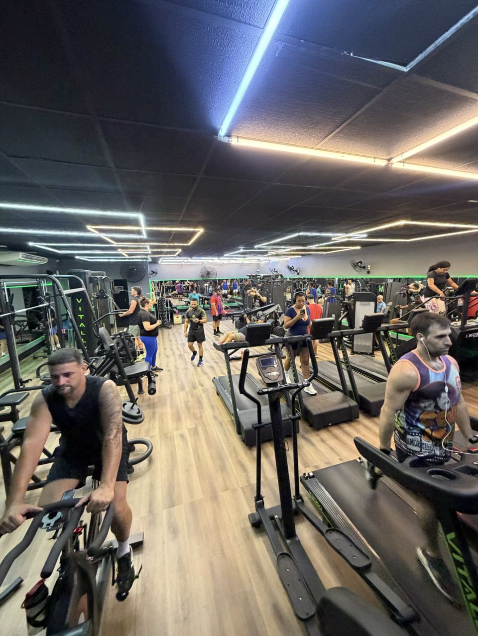 Sala ampla da Moraes Fit com bikes, esteiras e aparelhos, piso de madeira e linhas de luz no teto